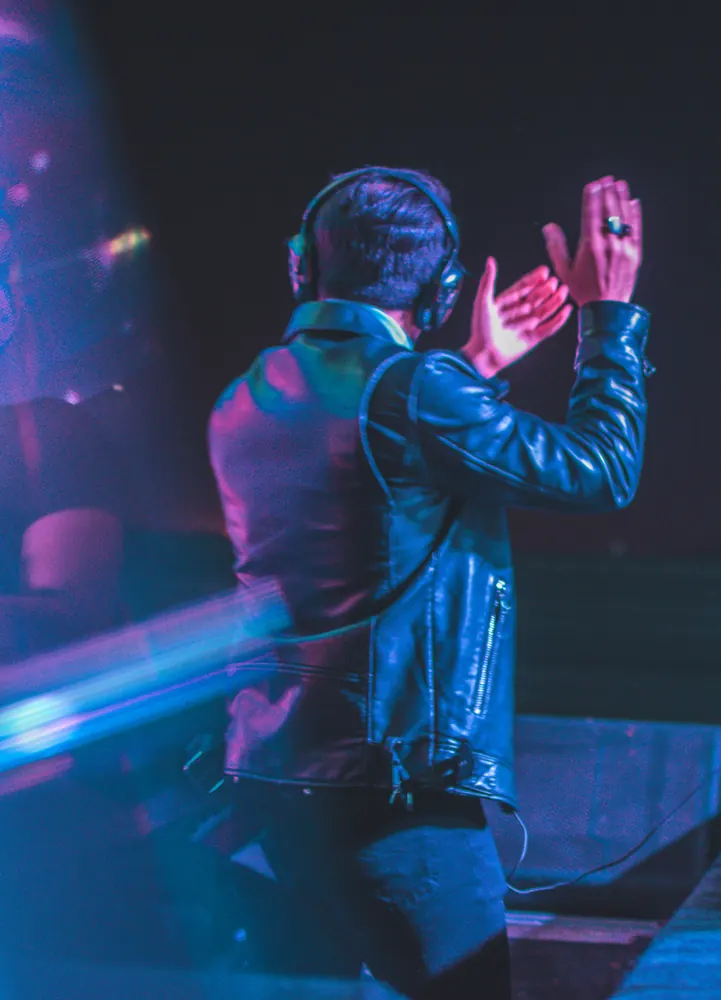 A DJ in headphones and a leather jacket, hands raised, in blue light