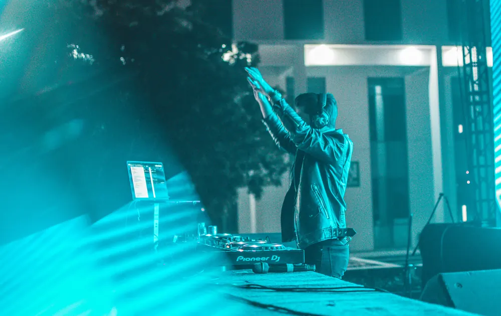 A DJ, arms up, behind the decks, in teal light