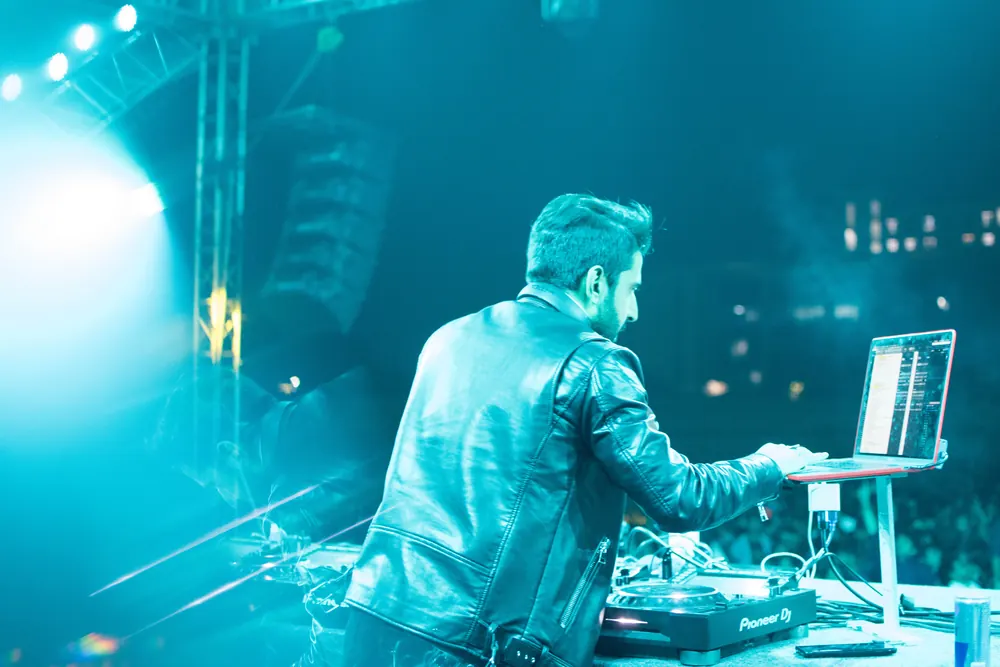 A DJ at the laptop, the lights of the crowd beyond, in teal
