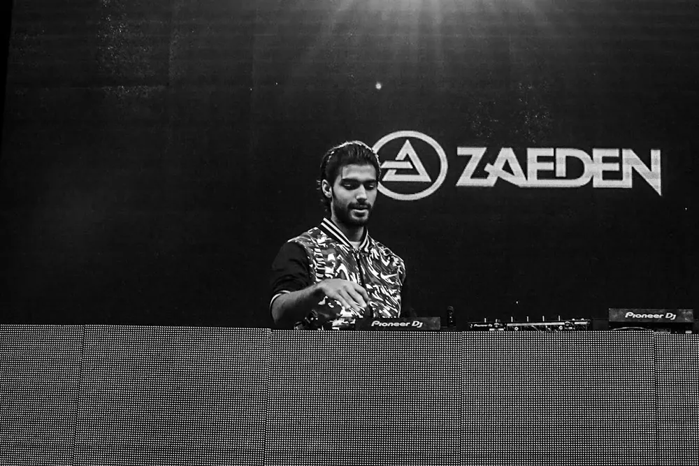 Zaeden at the decks, his name on the screen behind him, in black and white